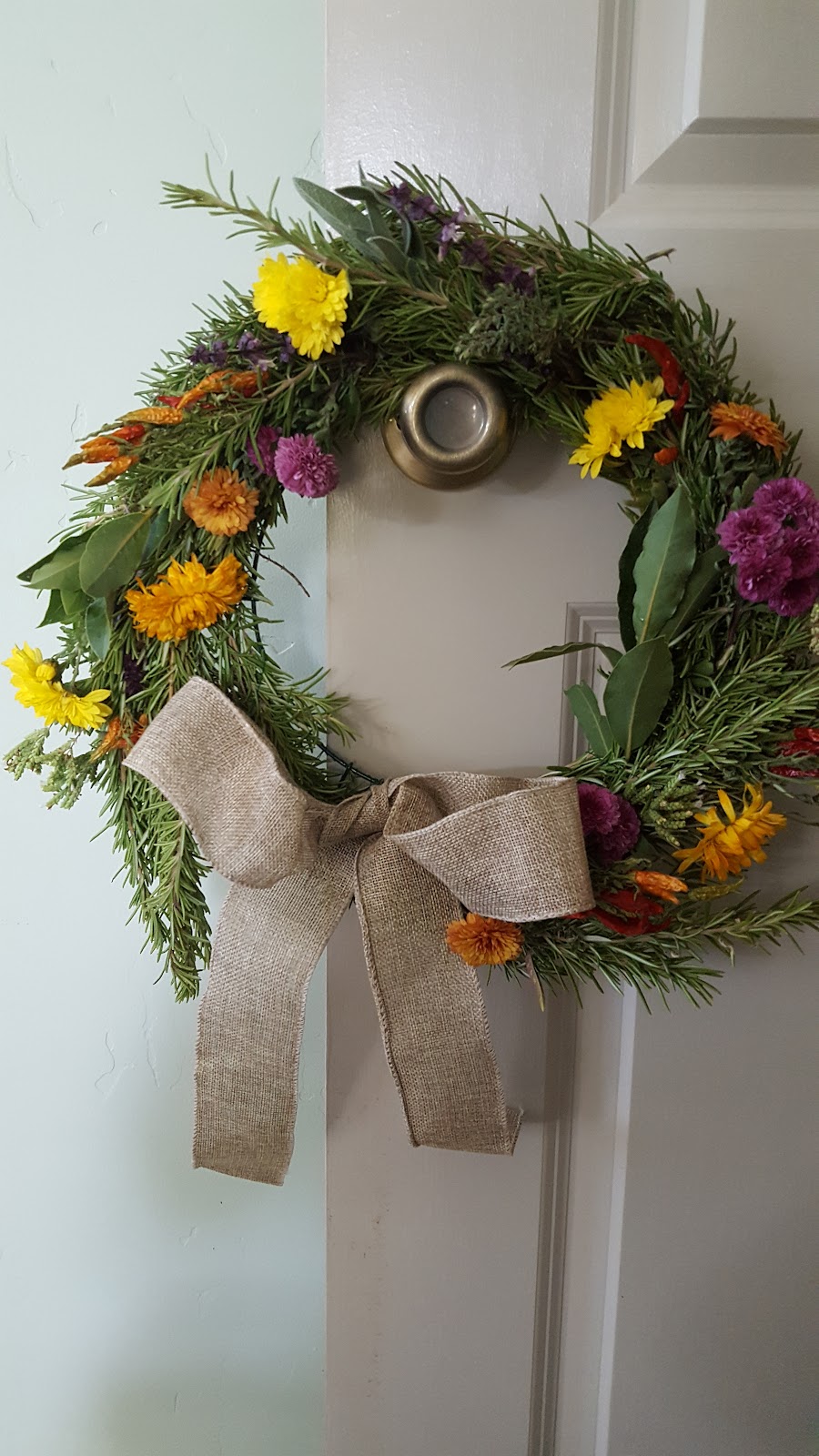 A handmade herbal wreath of rosemary and bay with dried flowers and a burlap bow, hung on a door